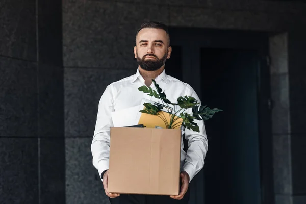 El hombre desempleado se muda del centro de oficinas con la caja llena de cosas personales, mirando tristemente a la cámara — Foto de Stock