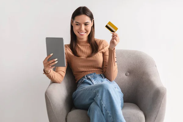 Весела молода леді з кредитною карткою і планшетним ПК сидить у кріслі, купує онлайн, використовуючи банківську програму — стокове фото
