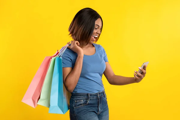 Donna africana con smartphone e borse della spesa in posa, sfondo giallo — Foto Stock