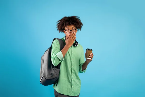 Cansado afroamericano estudiante masculino con café para ir y mochila bostezando en fondo de estudio azul — Foto de Stock