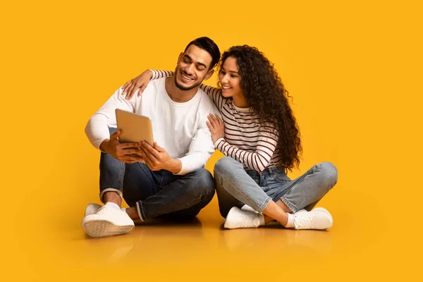 快乐的中东年轻夫妇坐在黄色的背景上，使用数码记事板 — 图库照片