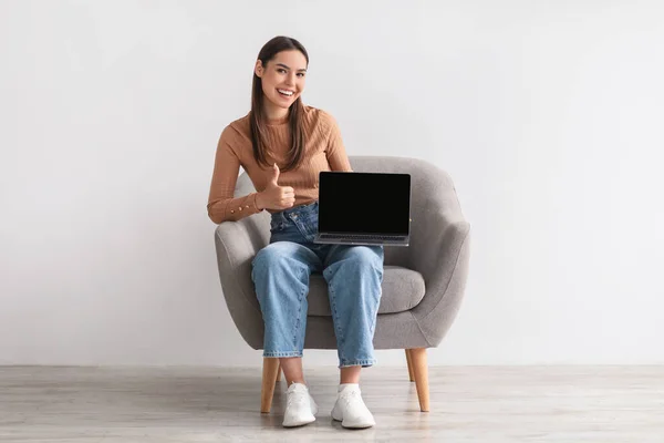 Feliz joven sentada en sillón, sosteniendo el ordenador portátil con la pantalla vacía y mostrando el pulgar hacia arriba, maqueta para el sitio web —  Fotos de Stock