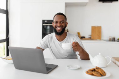 Afro-Amerikalı adam dizüstü bilgisayar kullanıyor kahve içiyor ve kameraya bakıyor.