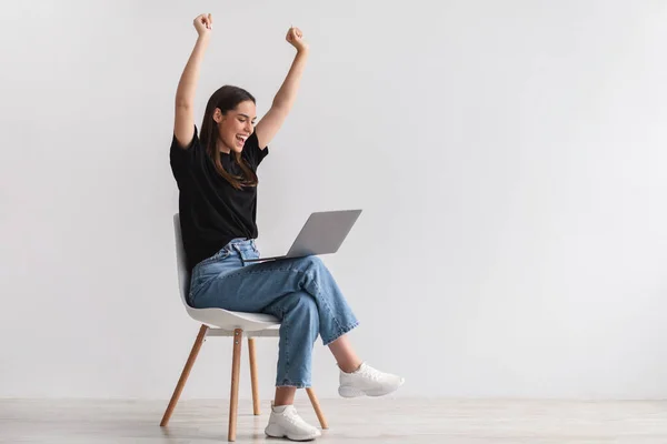 Nuori nainen istuu tuolilla kannettavan tietokoneen kanssa, nostaen kädet ylös, innoissaan lottovoitosta tai liiketoiminnan menestyksestä, kopioi tilaa — kuvapankkivalokuva