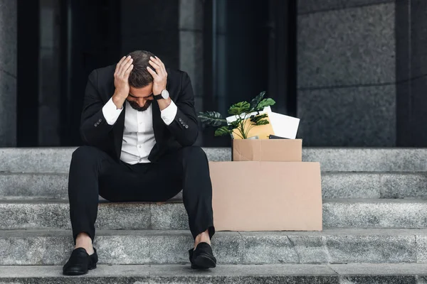 Upset mature businessman sitting with empty poster cardboard sign and box of personal stuff on stairs of office center — Stock Photo, Image