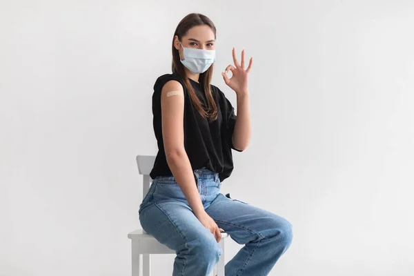 Giovane donna caucasica in maschera facciale dopo immunizzazione covid-19, mostrando spalla con cerotto, gesticolando bene — Foto Stock