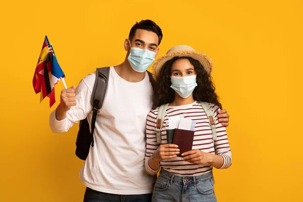 Viajes pandémicos. Pareja joven con máscaras médicas sosteniendo pasaportes y banderas internacionales —  Fotos de Stock