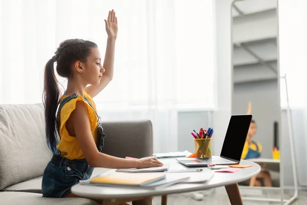 Menina sentada à mesa, usando laptop, levantando a mão para resposta — Fotografia de Stock