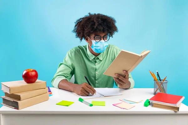 Black student guy wearing protective face mask and taking notes from book, sitting at table over blue background