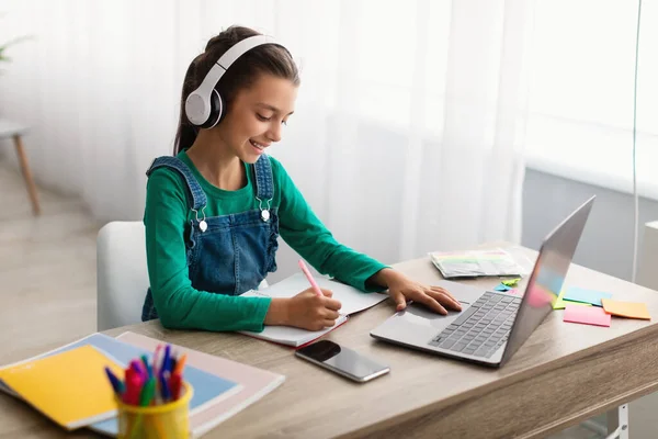 Дівчина сидить за столом, використовуючи ноутбук, пишучи в підручнику — стокове фото