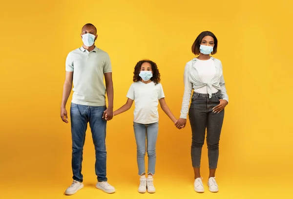 Jóvenes padres negros y su hija menor con máscaras faciales médicas y de la mano, fondo amarillo —  Fotos de Stock