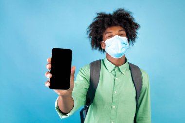 Koruyucu maskeli siyah bir öğrenci elinde boş ekranlı akıllı bir telefonla mavi arka planda duruyor.