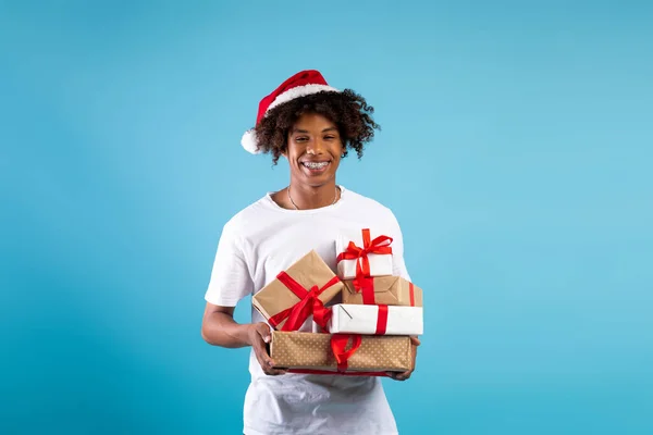 Time for celebration. Overjoyed black guy in santa hat holding stack of present boxes over blue background