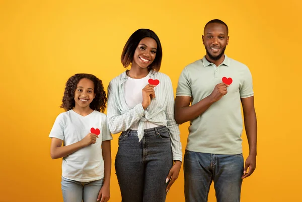 Adorável família afro-americana de três segurando cartões de papel vermelho em formas de coração, fundo amarelo — Fotografia de Stock