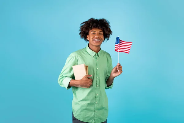 Échange d'étudiants et apprentissage de l'anglais. Zoomer heureux mec noir avec des livres et petit drapeau des États-Unis, fond bleu — Photo