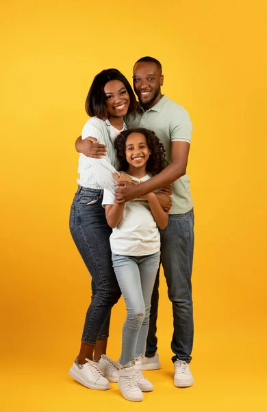 Egészséges családi kapcsolat. Afrikai amerikai apa és anya átölelő mosolygós lánya áll sárga háttér — Stock Fotó