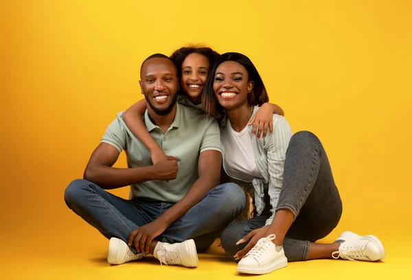 Retrato de família. Pais afro-americanos felizes e sua filha abraçando e sorrindo, sentados em fundo amarelo — Fotografia de Stock