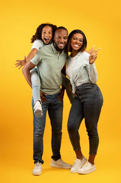 Vreugdevol Afrikaans Amerikaans gezin met leuke dochter poseren over gele achtergrond, plezier hebben, meisje meeliften vader — Stockfoto