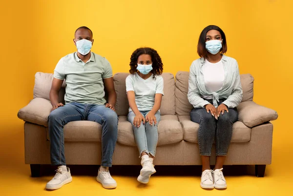 Joven familia afroamericana de tres, padres y su hija pequeña en máscaras protectoras sentados en el sofá —  Fotos de Stock