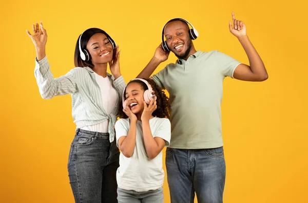 Desfrutem de música juntos. Feliz família afro-americana de três dançando em fones de ouvido, isolados em fundo amarelo — Fotografia de Stock