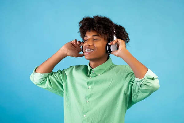 Улюблена пісня. Розслаблений афроамериканець у навушниках слухає музику закритими очима, стоїть на синьому фоні. — стокове фото