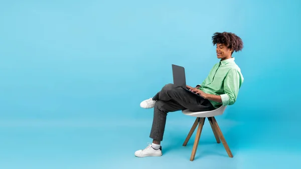 Koncepcja e-learningu. Afroamerykanin student facet za pomocą laptopa, siedzi na krześle na niebieskim tle, panorama, wolna przestrzeń — Zdjęcie stockowe