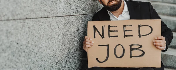 Necesito trabajo. Empresario sosteniendo cartel mientras está sentado al aire libre en las escaleras cerca del centro de oficina, espacio para copiar, cosecha, panorama — Foto de Stock