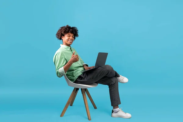 Heureux noir adolescent guy assis avec ordinateur portable dans chaise et montrant pouce vers le haut, recommander des cours en ligne, fond bleu — Photo