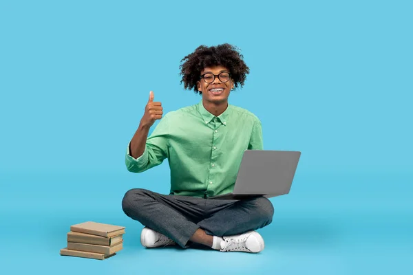 Heureux afro-américain adolescent guy assis sur le sol avec ordinateur portable et livres à proximité, montrant pouces vers le haut sur fond bleu — Photo