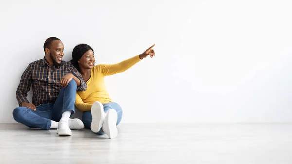 Czarne Millennial małżonkowie siedzi na podłodze w pobliżu białej ściany i wskazując obok — Zdjęcie stockowe