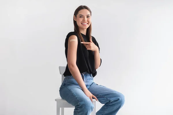 Feliz paciente joven apuntando a su brazo con curita después de la vacuna contra el coronavirus inyectada en el fondo del estudio de luz — Foto de Stock