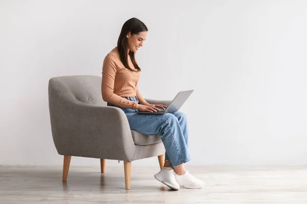 Jovencita feliz sentada en sillón, usando laptop, estudiando remotamente o teniendo reuniones de negocios, espacio libre —  Fotos de Stock