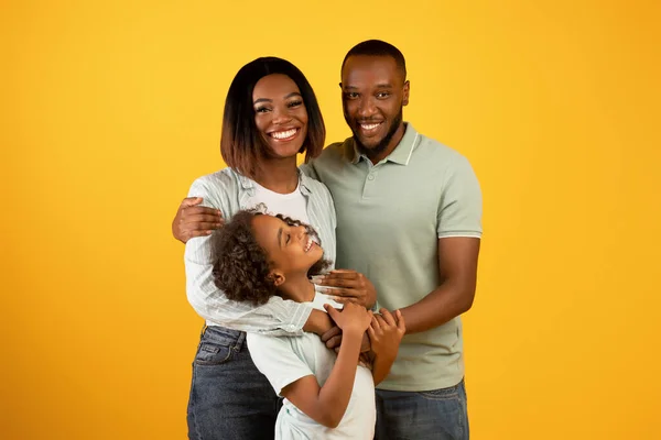 Familia feliz. Amantes padres afroamericanos con hija abrazando y sonriendo a la cámara sobre fondo amarillo — Foto de Stock