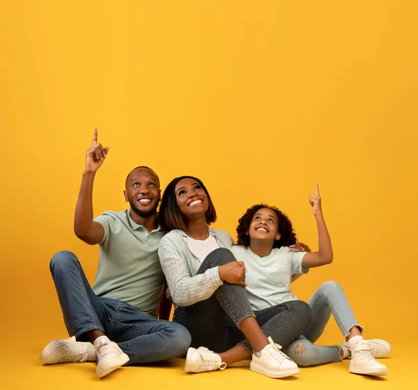 Mira ahí. Familia afroamericana feliz señalando los dedos hacia arriba en el espacio vacío, sentado en el suelo sobre el fondo amarillo —  Fotos de Stock