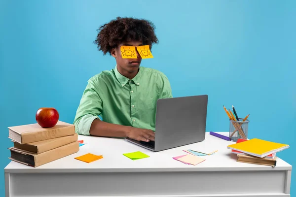 Agotado adolescente con los ojos pintados en pegatinas sentado en el escritorio usando el ordenador portátil, durmiendo sobre fondo azul — Foto de Stock