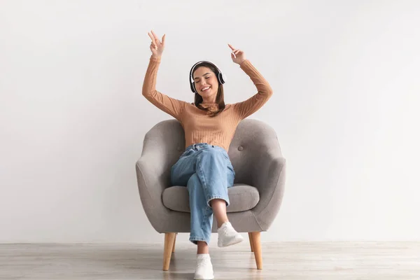 Jovencita guay sentada en sillón, escuchando música en auriculares inalámbricos, bailando su canción favorita — Foto de Stock