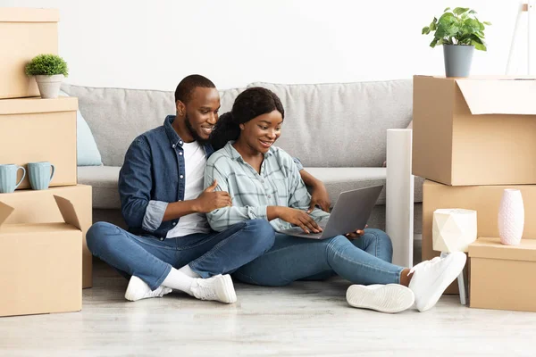 Glimlachende zwarte stelt zittend met laptop tussen kartonnen dozen in nieuw huis — Stockfoto