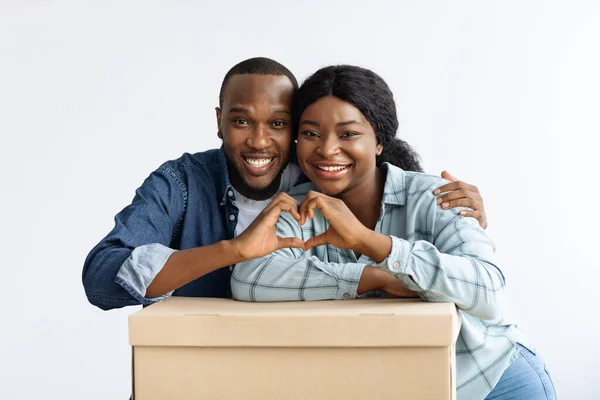 Bienes raíces. Cónyuges negros felices apoyados en la caja de cartón, haciendo el gesto del corazón —  Fotos de Stock