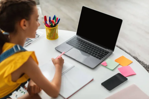 Chica sentada en el escritorio, usando un portátil en blanco, escribiendo en un libro de texto —  Fotos de Stock