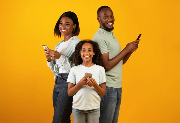 Familia moderna. Emocionado padre afroamericano, madre e hija usando teléfonos inteligentes sobre fondo amarillo — Foto de Stock