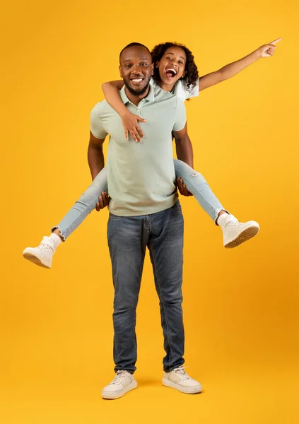 Os pais adoram. Saindo pai negro carregando sua filha nas costas, se divertindo e passando tempo juntos no fundo amarelo — Fotografia de Stock