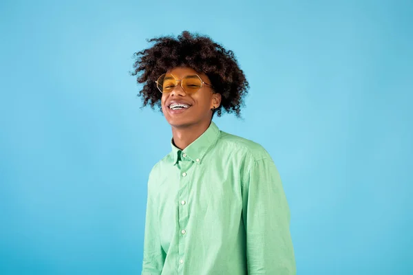 Retrato de feliz afro-americano adolescente cara em camisa de hortelã e óculos de sol amarelos posando no fundo do estúdio azul — Fotografia de Stock