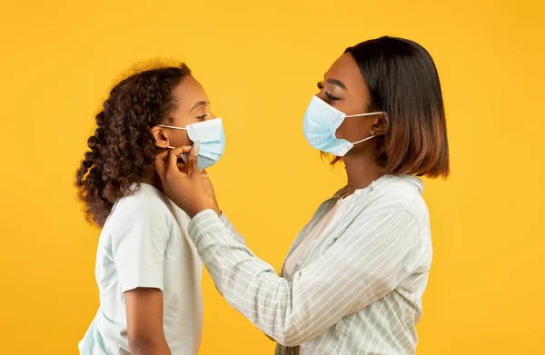 Amoroso afro-americano mulher colocando máscara médica em seu rosto bonito filha, fundo estúdio amarelo — Fotografia de Stock