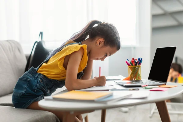 女孩坐在办公桌前，使用空白笔记本电脑，用课本写作 — 图库照片