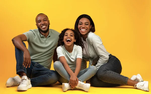 Gelukkig afrikaanse amerikaanse familie zitten op de vloer en lachen, samen glimlachen naar camera over gele studio achtergrond — Stockfoto