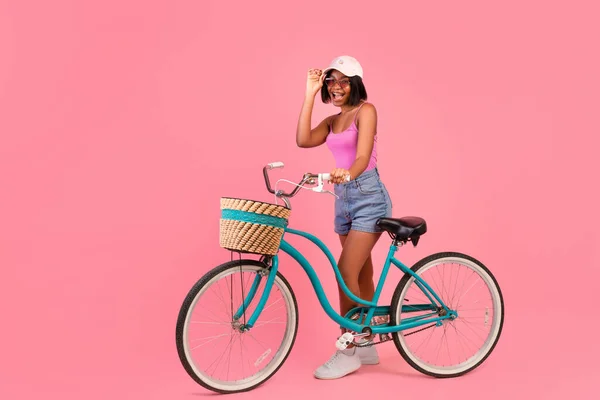 Full length portrait of emotional black lady in summer clothes standing with bike on pink studio background, copy space — Stock Photo, Image