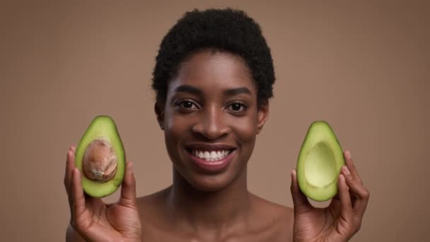 Mujer afroamericana sosteniendo mitades de aguacate cerca de la cara, fondo beige — Vídeos de Stock