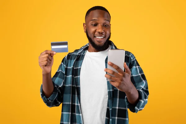 Happy African Mężczyzna gospodarstwa karty kredytowej i telefonu, żółte tło — Zdjęcie stockowe