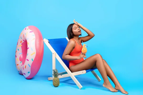 Comprimento total de jovem negra sofrendo de calor de verão, segurando coquetel tropical enquanto sentada na cadeira lounge — Fotografia de Stock