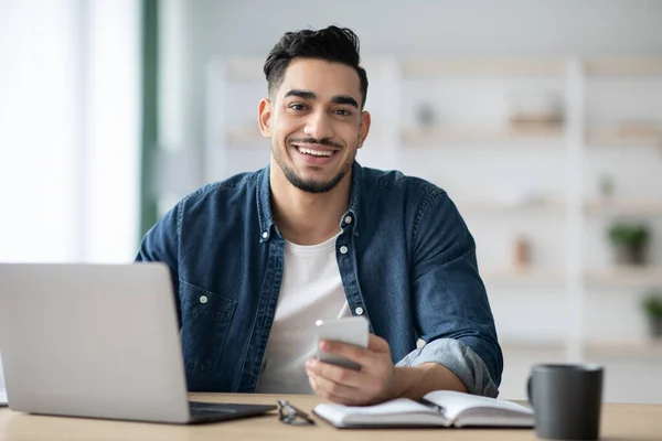 Usmívající se arabsky chlap pomocí smartphone zatímco sedí na workkdesk — Stock fotografie